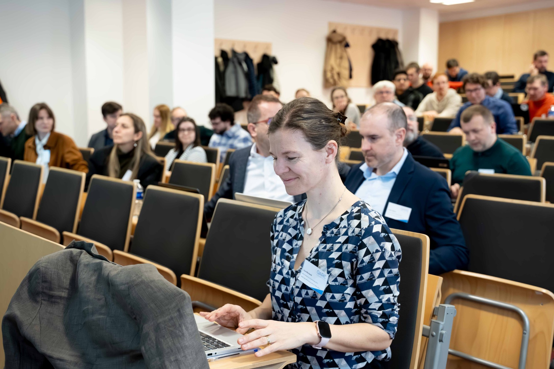 Konferences auditorijā sieviete sēž rindā un strādā ar klēpjdatoru, fonā citi dalībnieki.