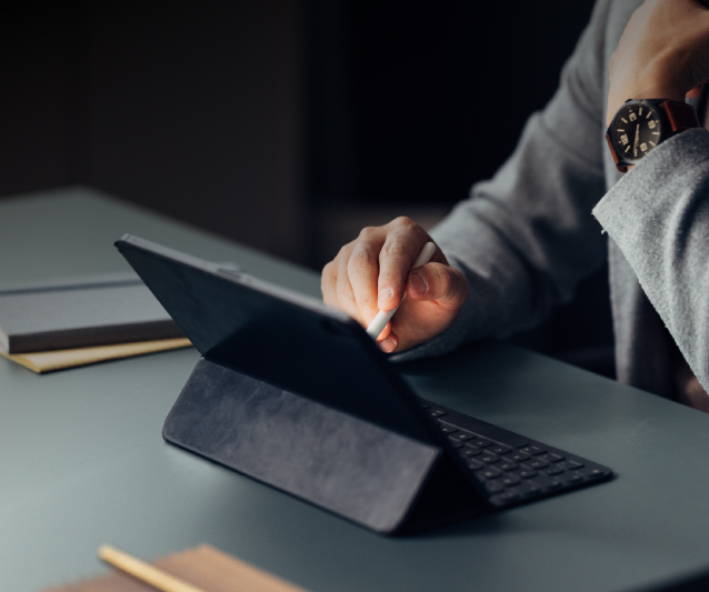 Roka ar planšetdatora pildspalvu raksta planšetdatorā ar pieslēgtu tastatūru uz galda.