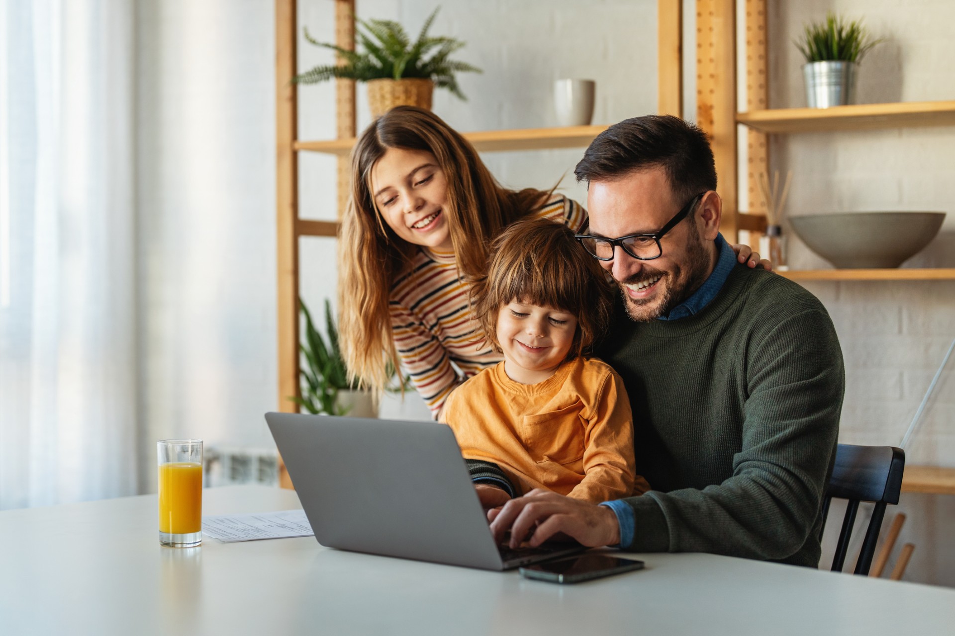 Tēvs ar bērnu klēpī un māte kopā skatās klēpjdatorā mājās; uz galda glāze sulas.