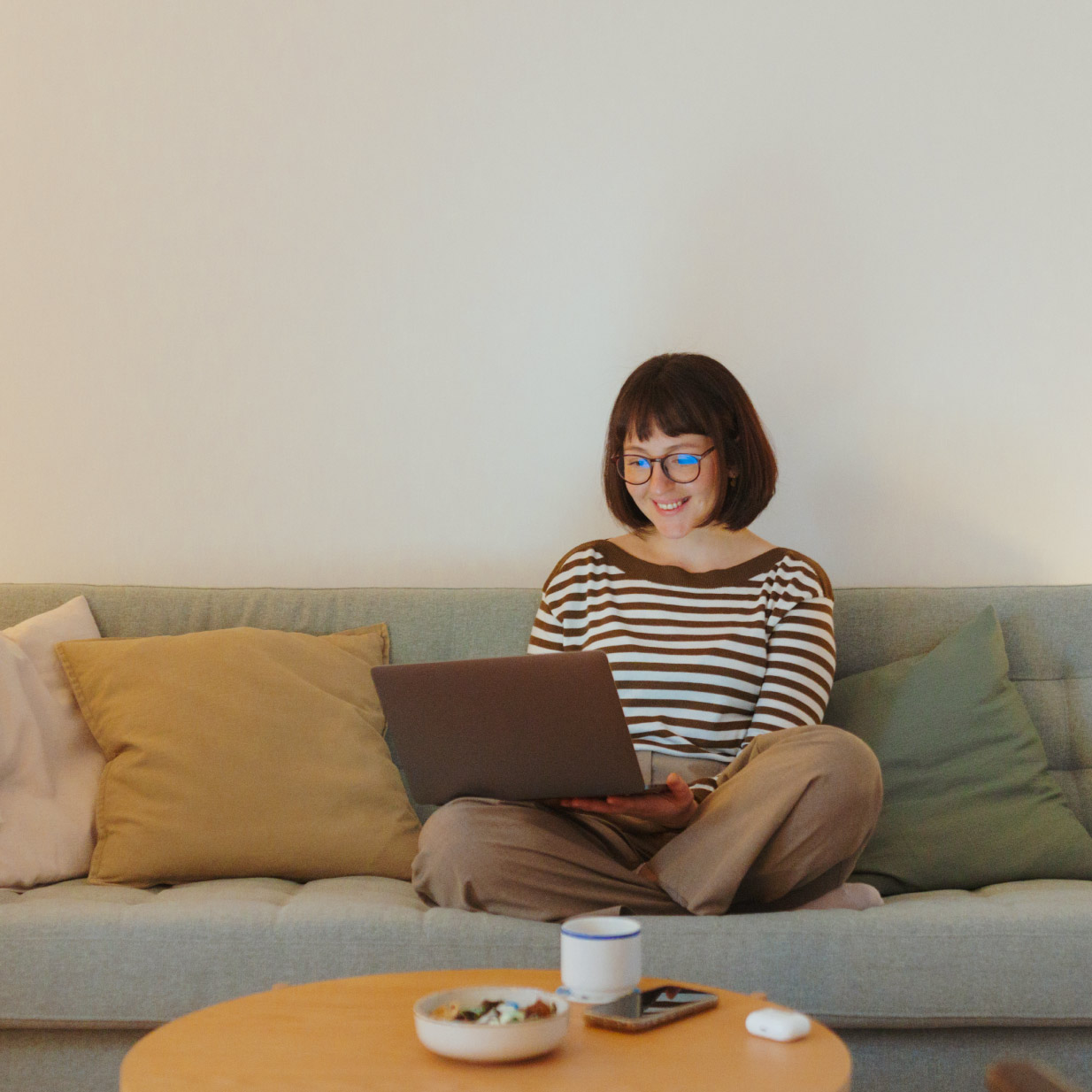 Sieviete sēž uz di'vana, smaida un skatās portatīvajā datorā