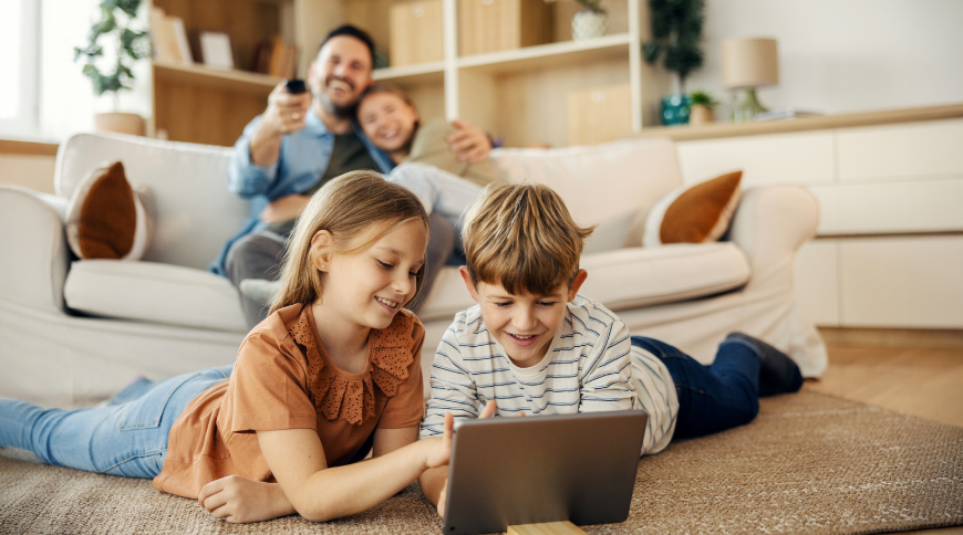 Bērni - puika un meitene, skatās planešetdatorā, fonā vecāki laimīgi skatās televīziju 