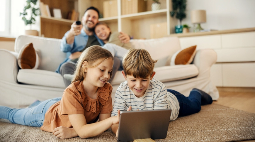 Bērni - puika un meitene, skatās planešetdatorā, fonā vecāki laimīgi skatās televīziju 