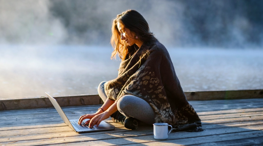 Meitene pie ezera, uz laipas, skatās portatīvajā datorā 