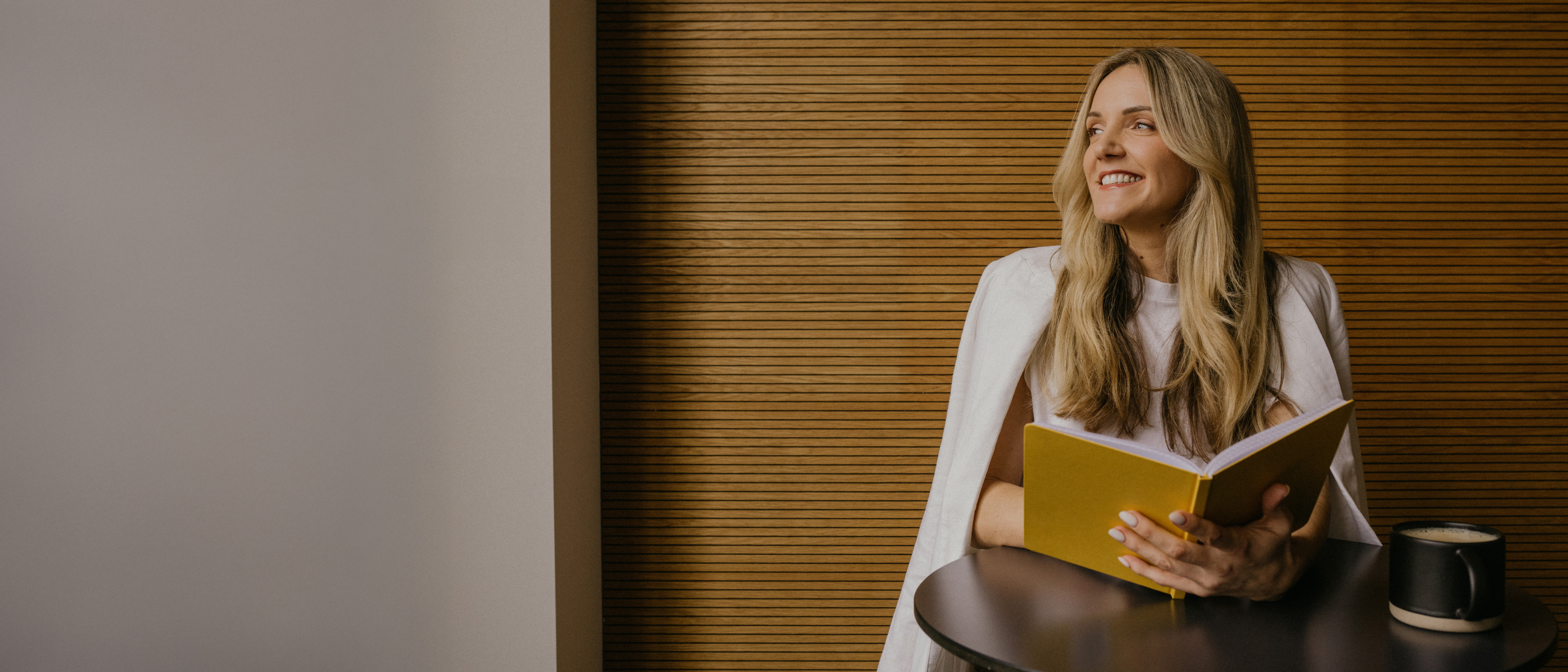  Smaidoša sieviete sēž pie galdiņa un lasa dzeltenu grāmatu; fonā koka lamelu siena, uz galda melna krūze.