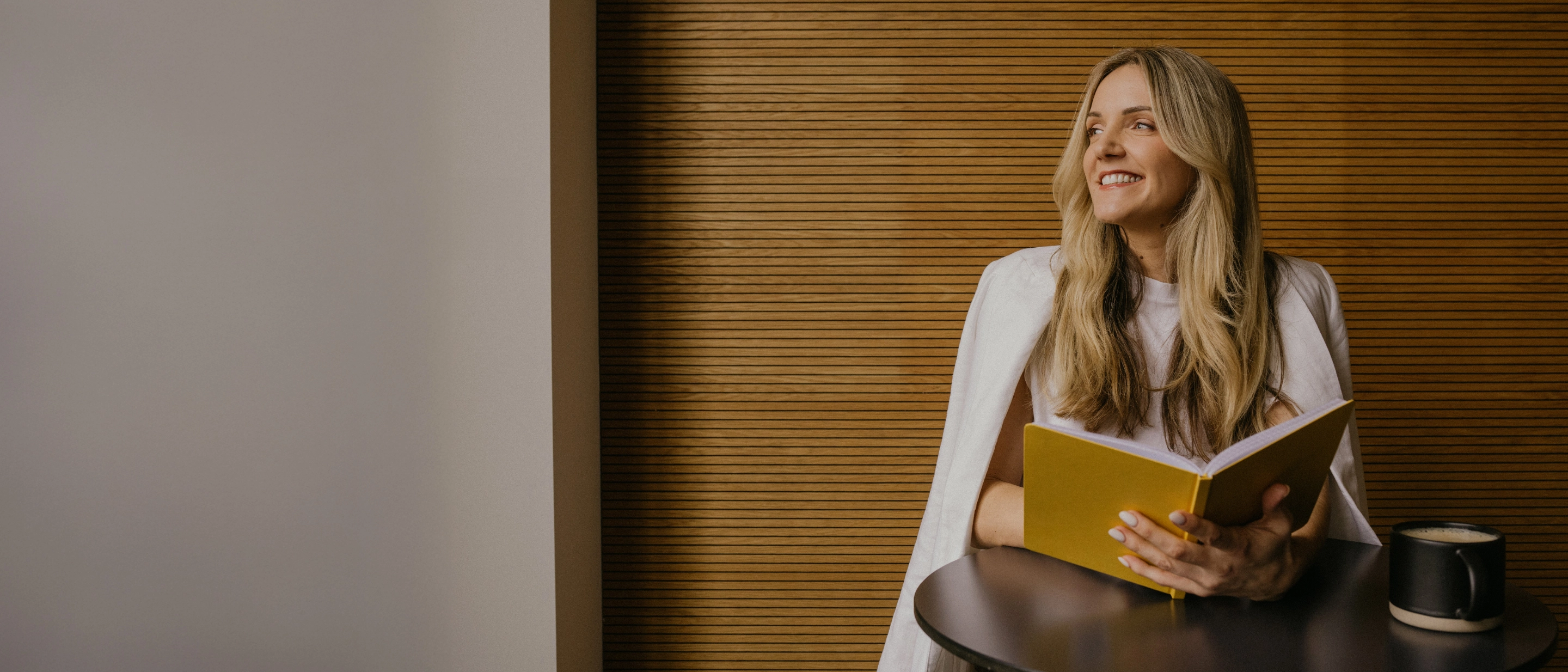  Smaidoša sieviete sēž pie galdiņa un lasa dzeltenu grāmatu; fonā koka lamelu siena, uz galda melna krūze.
