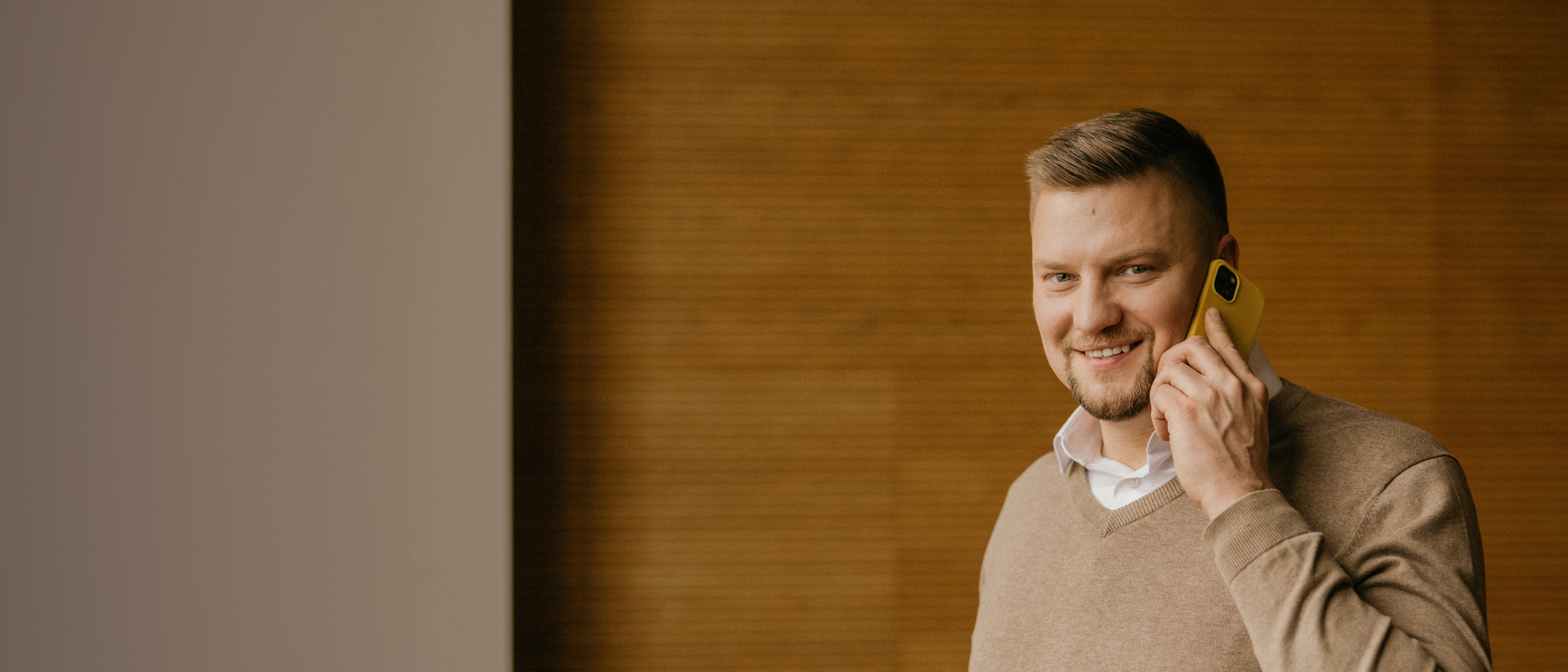 Vīrietis ar gaiši brūniem matiem un gaišā džemperī modernā birojā runā pa telefonu