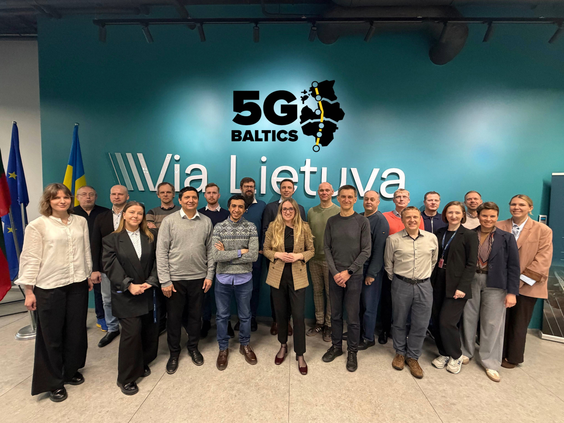 Grupas foto modernā konferenču telpā ar vairākām personām, kas stāv rindā pie sienas ar uzrakstiem '5G Baltics' un 'Via Lietuva'; fonā karogi un pasākuma vizuālais noformējums.