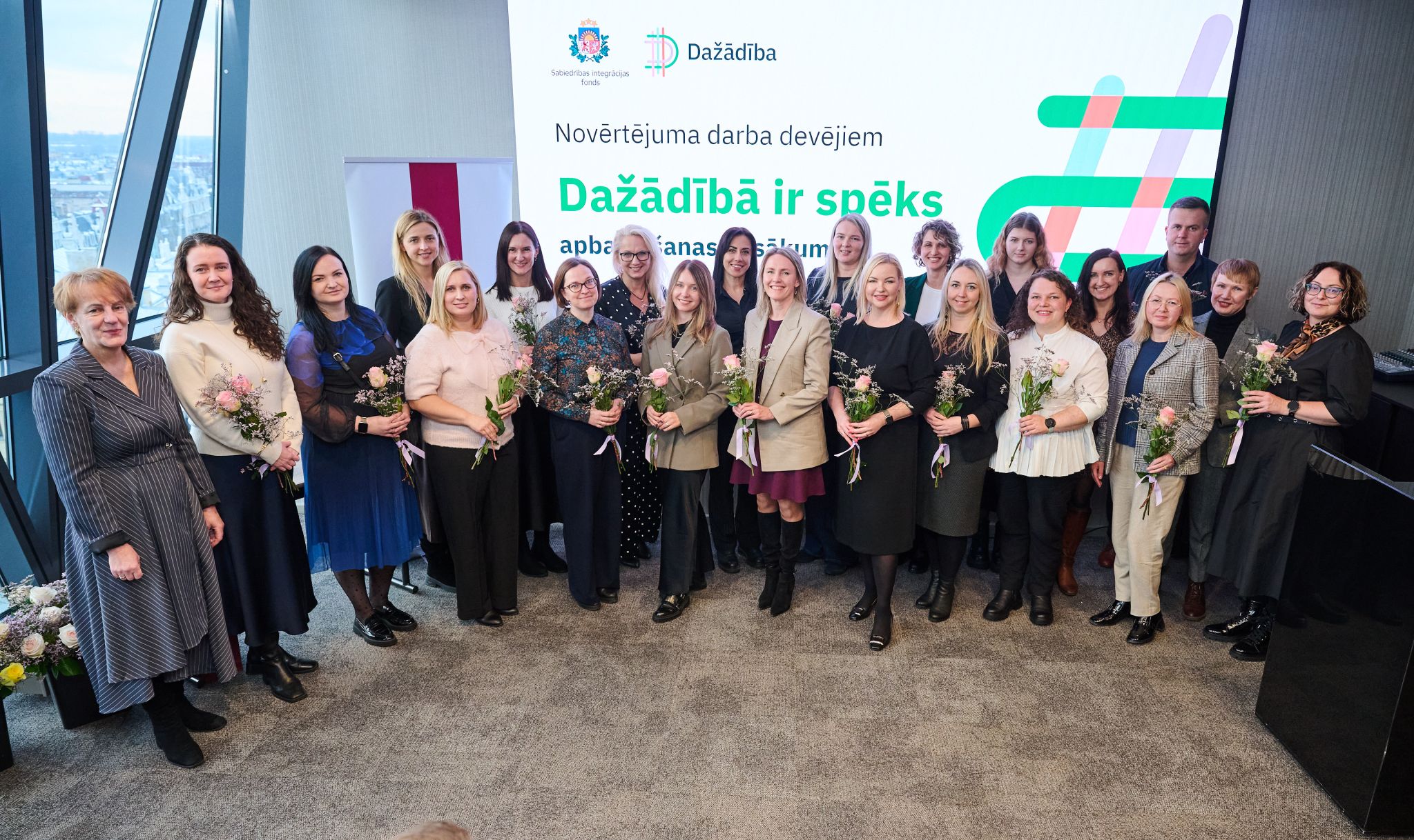 Kopbilde apbalvošanas pasākumā “Dažādībā ir spēks” — cilvēki ar ziediem stāv pie prezentācijas ekrāna.