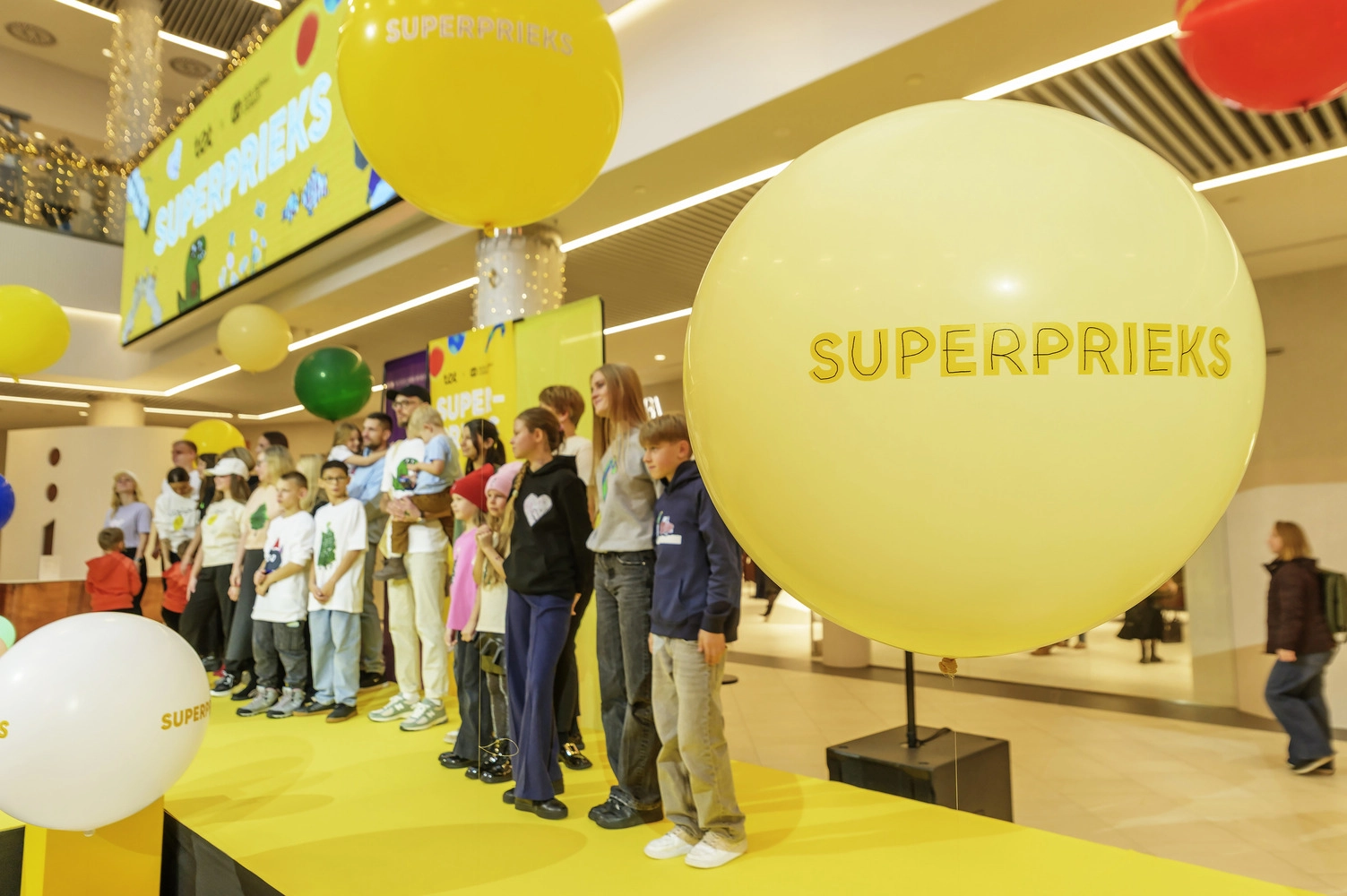 Cilvēku grupa uz dzeltenas skatuves pasākumā, ko ieskauj lieli dzelteni baloni ar uzrakstu “SUPERPRIEKS”.
