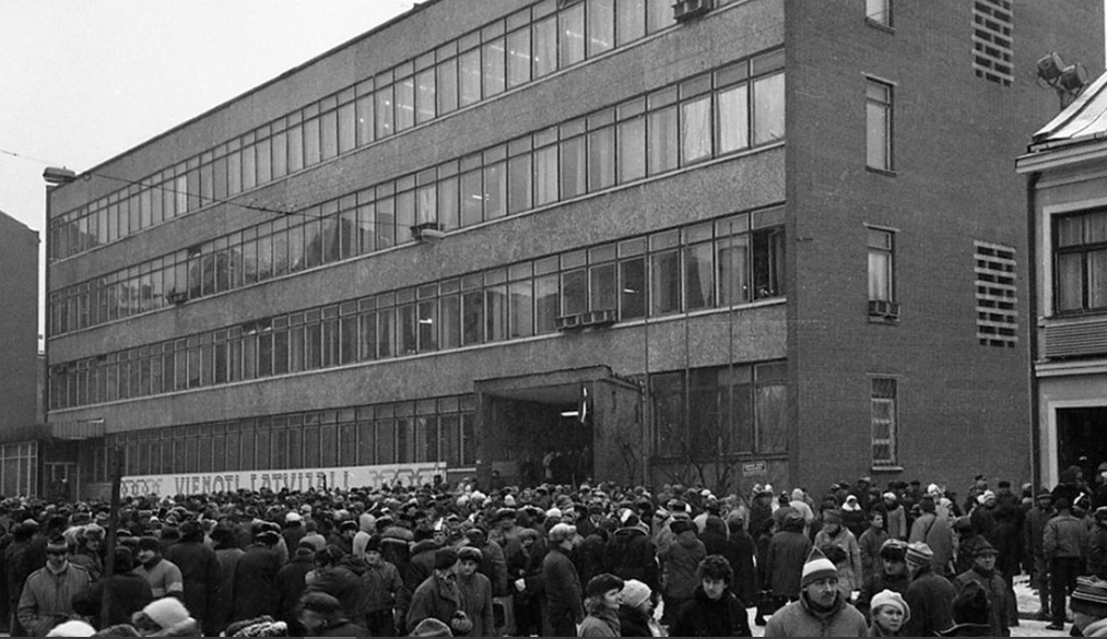 Liels cilvēku pūlis ziemas apģērbā stāv pie daudzstāvu ēkas. Attēls ir melnbalts, ēka izskatās pēc administratīvās ēkas, priekšā redzama ieeja ar nojumi.