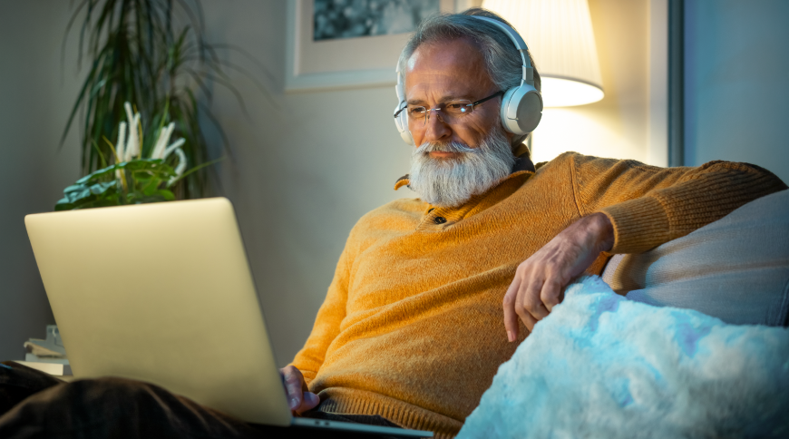 Vecs vīrs skatās portatīvajā datorā un klausās austiņas