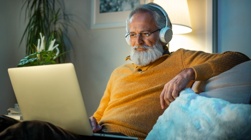 Vecs vīrs skatās portatīvajā datorā un klausās austiņas