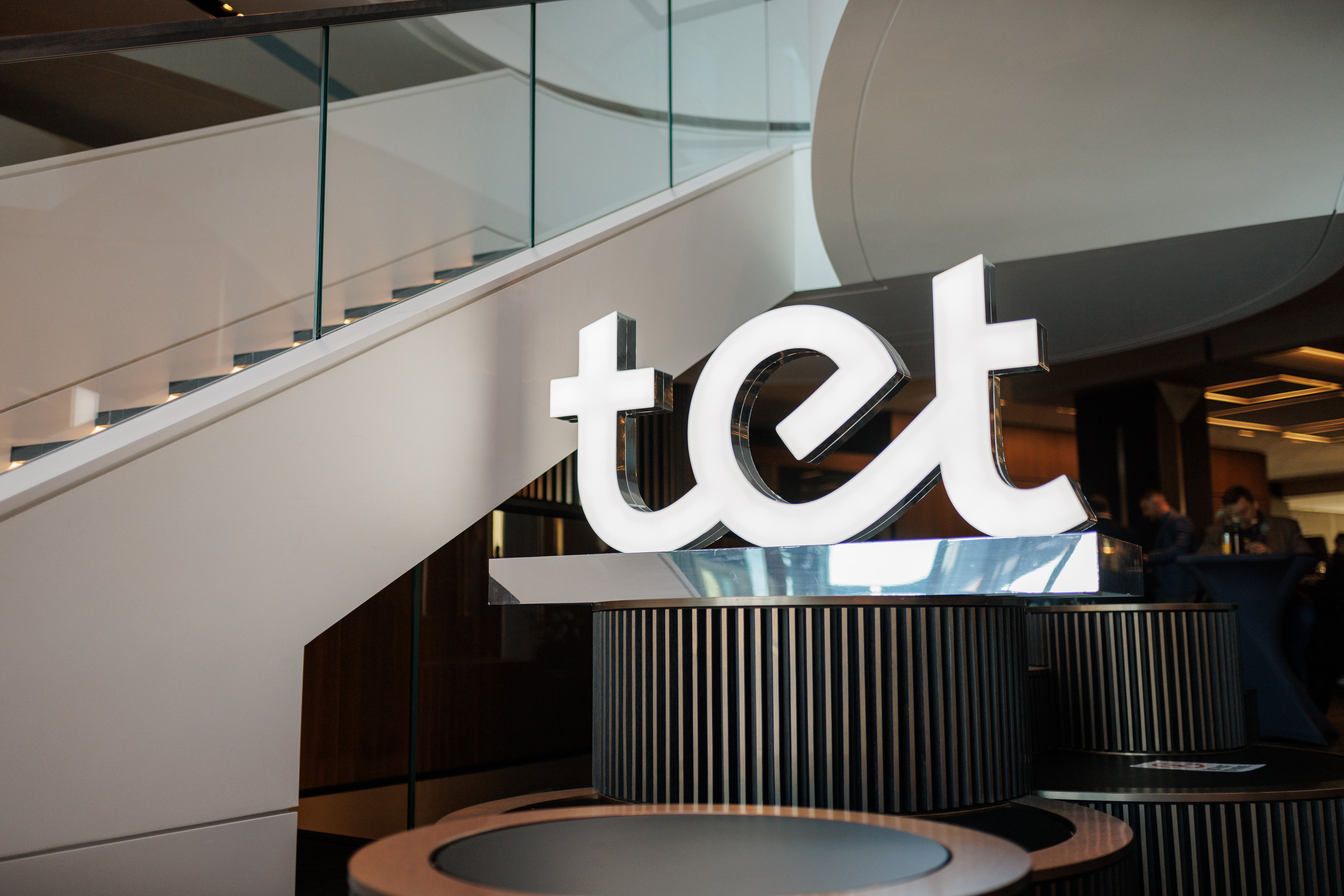 Izgaismots ‘tet’ logotips modernā interjerā ar līkumotām kāpnēm fonā.