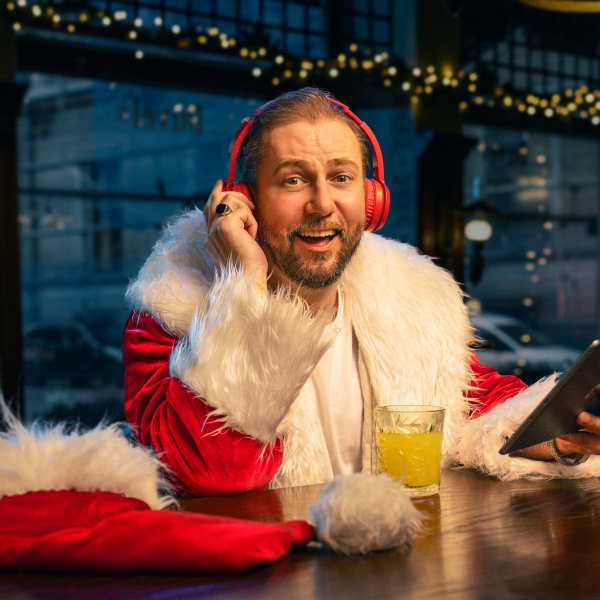 Vīrietis tērpies Ziemassvētku vecīša kostīmā ar baltām kažokādas apmalēm sēž pie galda ar planšetdatoru rokās un sarkanām austiņām ap ausīm. Uz galda atrodas dzeltens dzēriens stikla glāzē. Fonā svētku gaismas un dekorācijas.