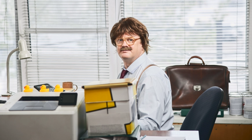 Vīrietis biroja vidē sēž pie galda ar dokumentu kaudzēm, mapēm un biroja tehniku, fonā logs ar žalūzijām un ādas portfelis uz skapīša.