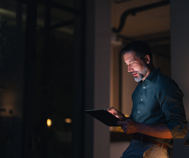 Skats uz konferenču telpu caur stikla sienu, kur vīrietis stāv pie galda ar planšetdatoru, bet trīs dalībnieki sēž krēslos un klausās runātājā.