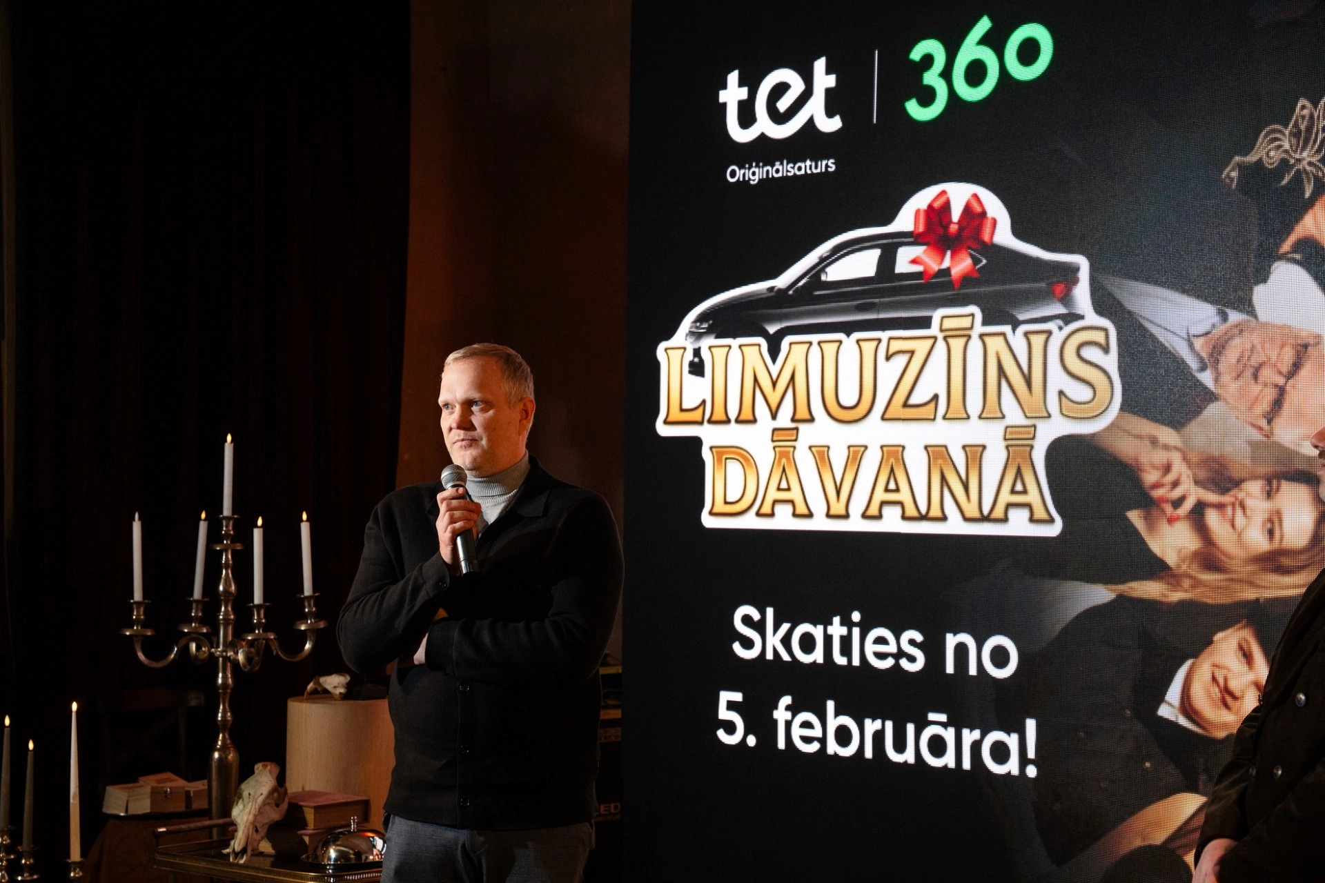 Vīrietis pelēkā džemperī runā mikrofonā Tet oriģinālsatura 'Limuzīns dāvanā' prezentācijas pasākumā. Fonā redzams seriāla plakāts ar tekstu 'Skaties no 5. februāra!' un svečturi interjerā.