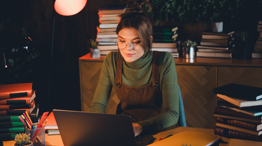 Persona ar brillēm strādā pie klēpjdatora; fonā grāmatu kaudzes un silts apgaismojums.