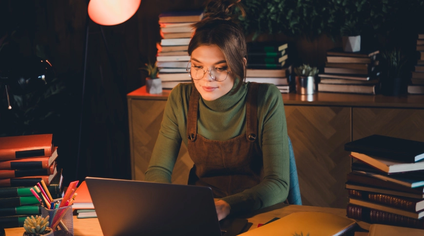 Persona ar brillēm strādā pie klēpjdatora; fonā grāmatu kaudzes un silts apgaismojums.