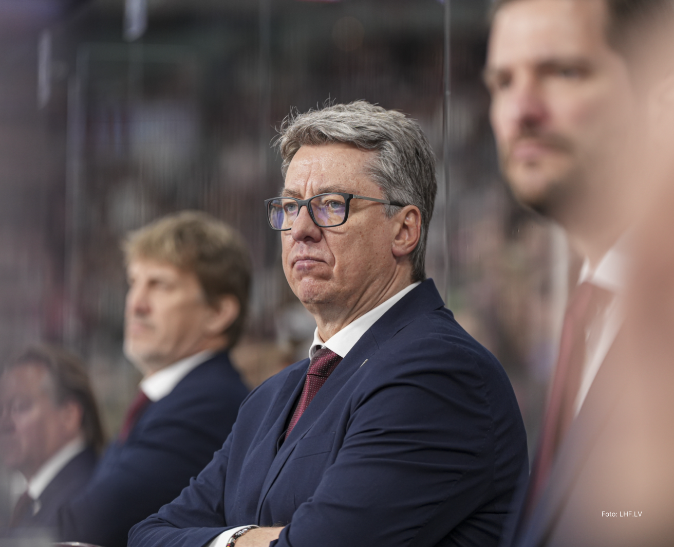 Tuvplāna kadrs ar Latvijas hokeja izlases treneri Hariju Vītoliņu sporta arēnas komandas zonā, sēžot aiz aizsargstikla; attēlā cilvēks tumšā uzvalkā ar kaklasaiti un brillēm, fonā izplūdušas citas personas un arēnas interjers ar skatītājiem.