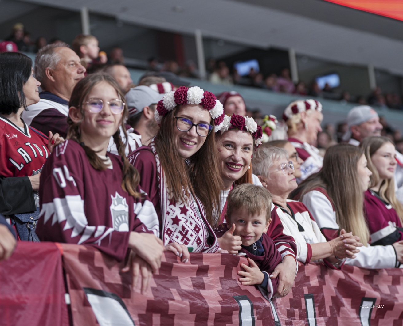 Hokeja līdzjutēju grupa sporta arēnā, tribīnēs cilvēki Latvijas karoga krāsas sporta kreklos ar Latvijai raksturīgiem etnogrāfiskiem rakstiem, daļai līdzjutēju galvās ziedu vainagi; visi sēž cieši blakus, priekšplānā bērns.