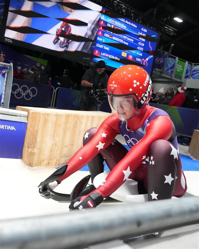Kamaniņu braucēja Elīna Ieva Bota sēž uzkamaniņām tikko pēc veiktā brauciena. Fonā olimpiskā simbolika, tablona ar rezultātiem, ekrāns un atbalstītāji.