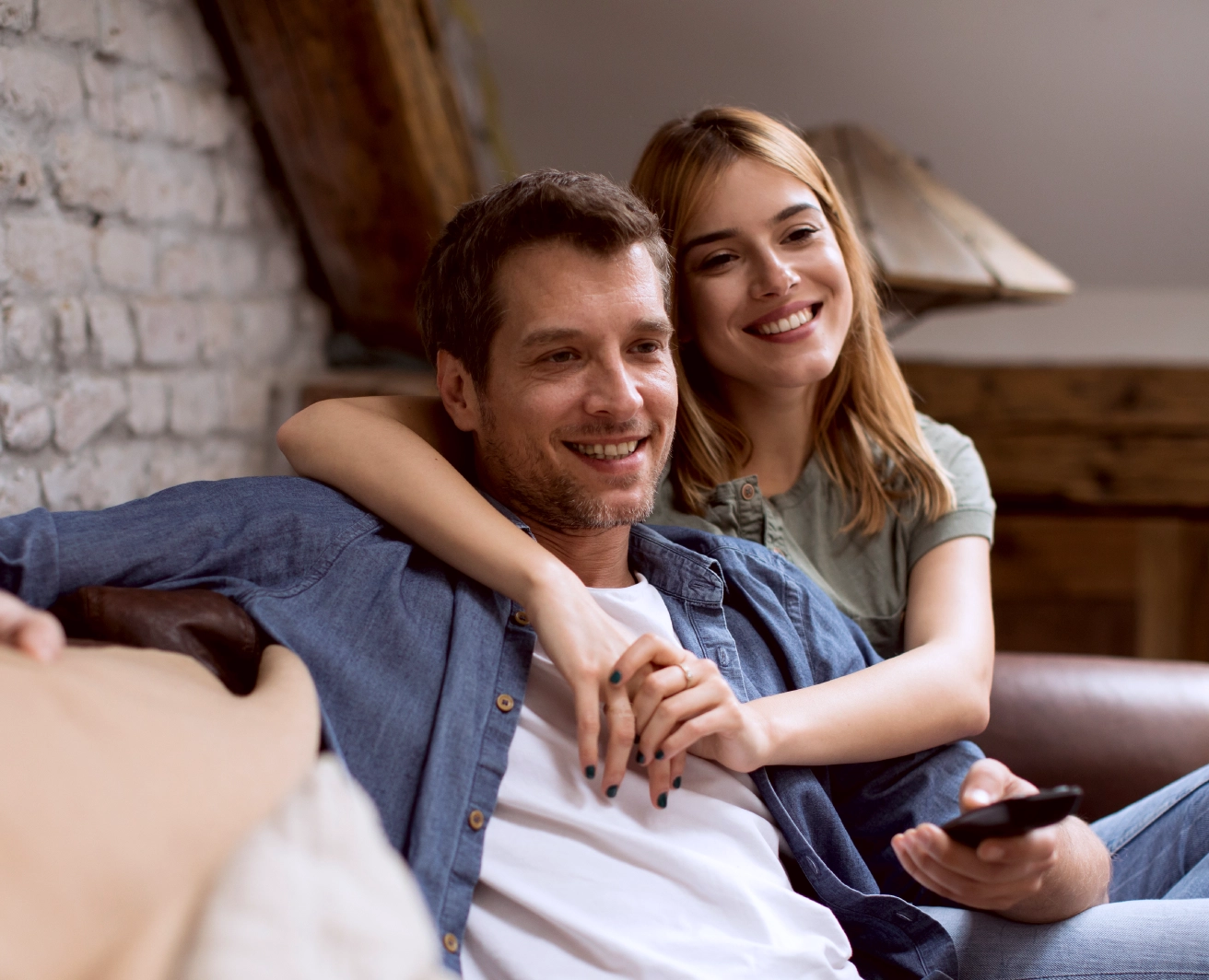 Vīrietis un sieviete sēž uz dīvāna mājīgā istabā, vīrietis tur pulti, bet sieviete ir apķērusi vīrieti ap pleciem, fonā redzama ķieģeļu siena un koka interjera elementi.