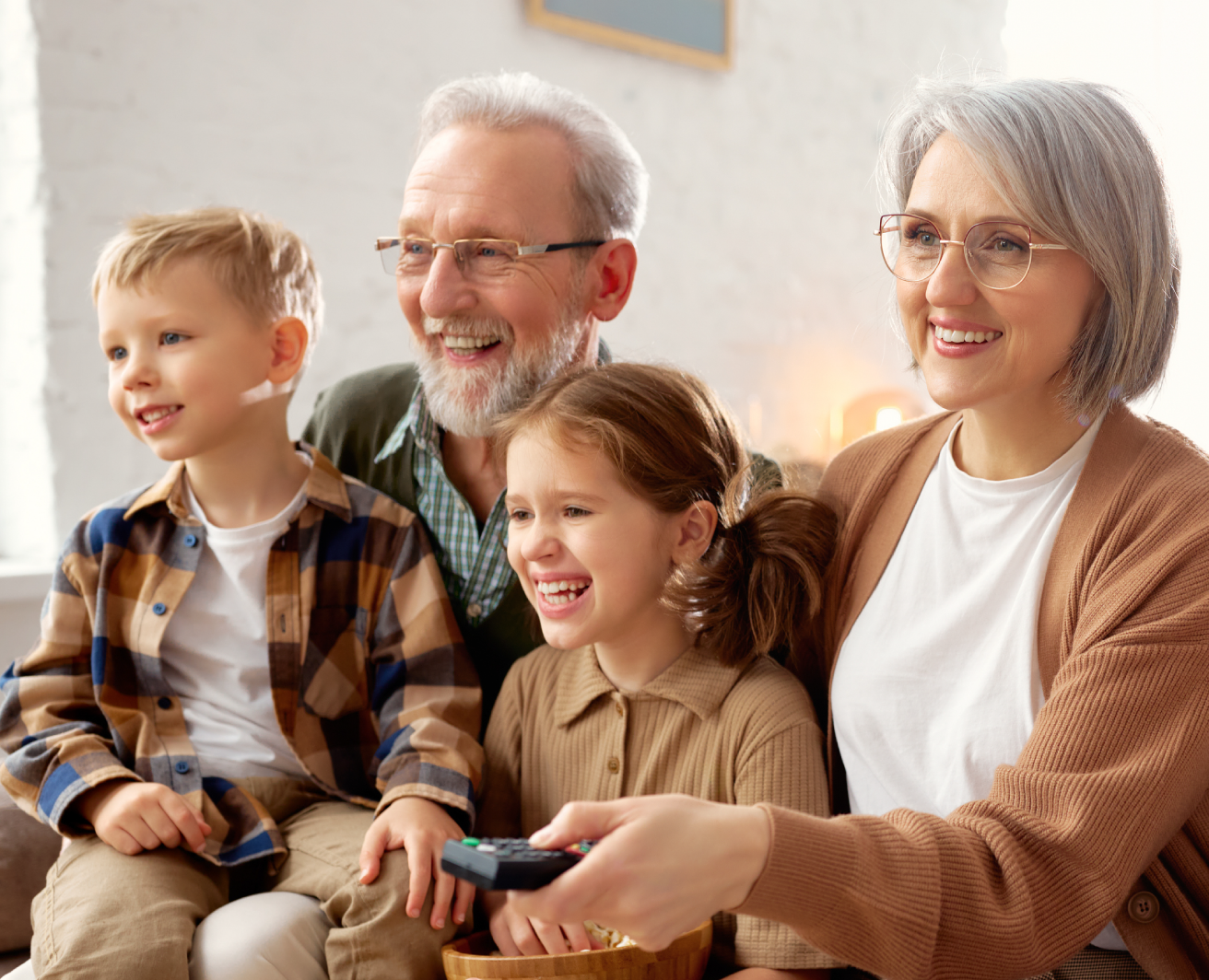 Četri cilvēki, vīrietis, sieviete un 2 bērni, sēž uz dīvāna mājīgā istabā. Sieviete rokā tur TV pulti, bērns koka tekstūras bļodu. Fonā silts apgaismojums.