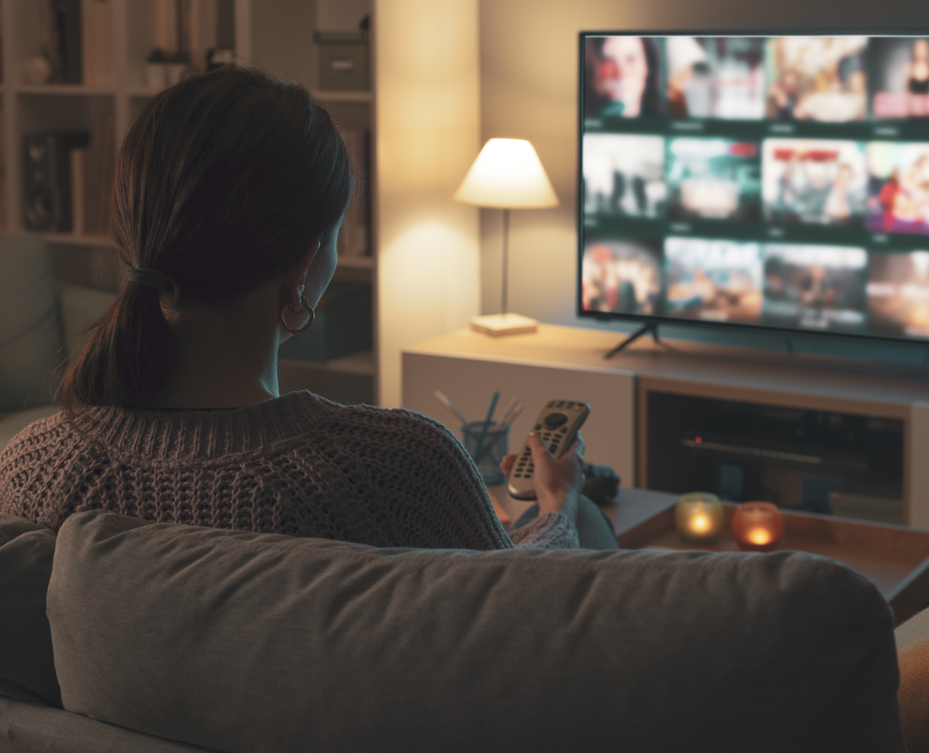Sieviete sēž uz dīvāna mājīgā istabā, skatās televizoru un tur TV pulti rokā. Televizora ekrānā straumēšanas platformas izvēlnes skats ar vairākām filmu un seriālu sīktēliem. Fonā galds ar iedegtām svecēm un galda lampu, kas rada siltu apgaismojumu.