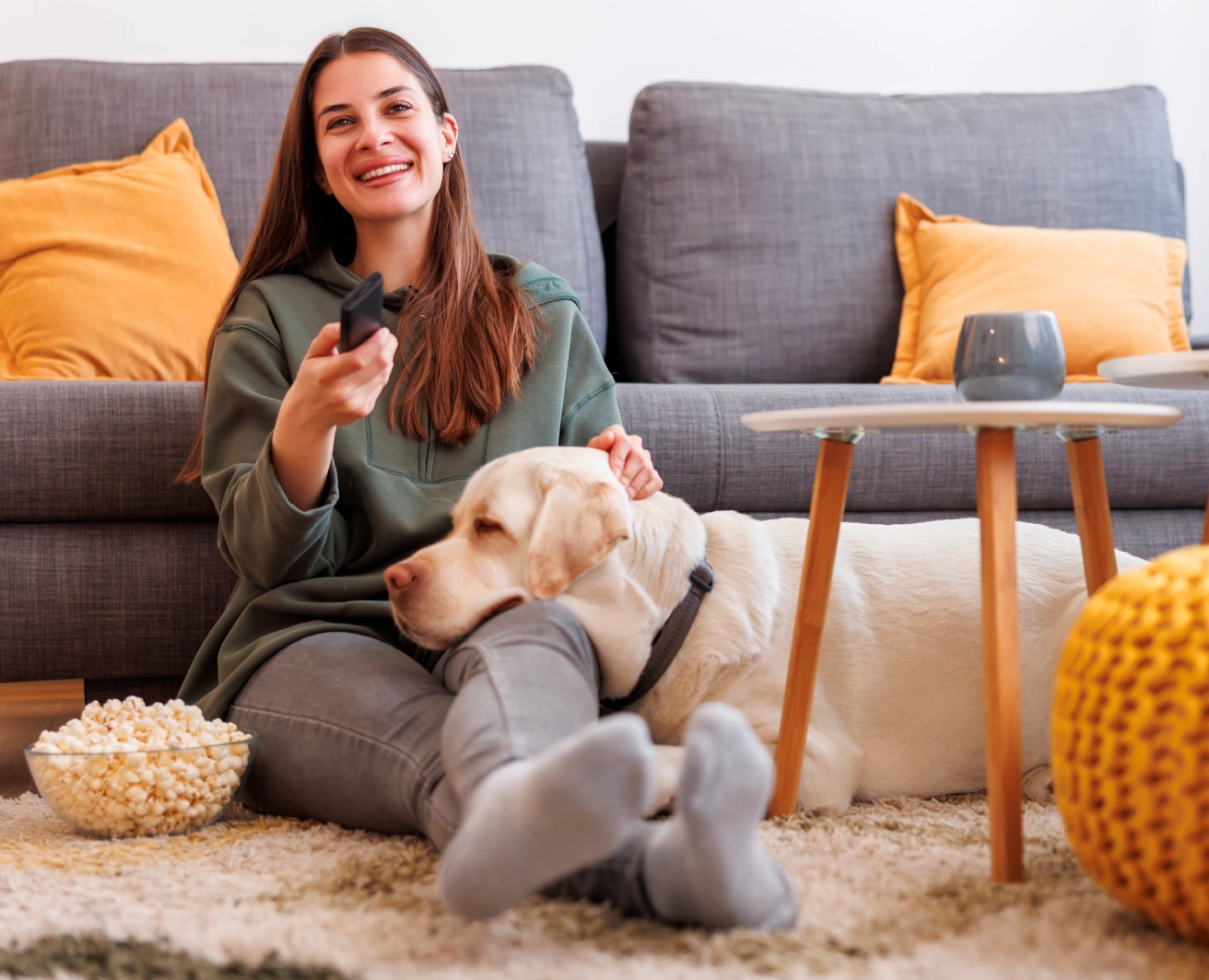 Sieviete sēž uz grīdas pie pelēka dīvāna, turot TV pulti vienā rokā un glāstot gaišu suni, kas ielicis savu galvu viņai klēpī. Uz grīdas ir mīksts paklājs.