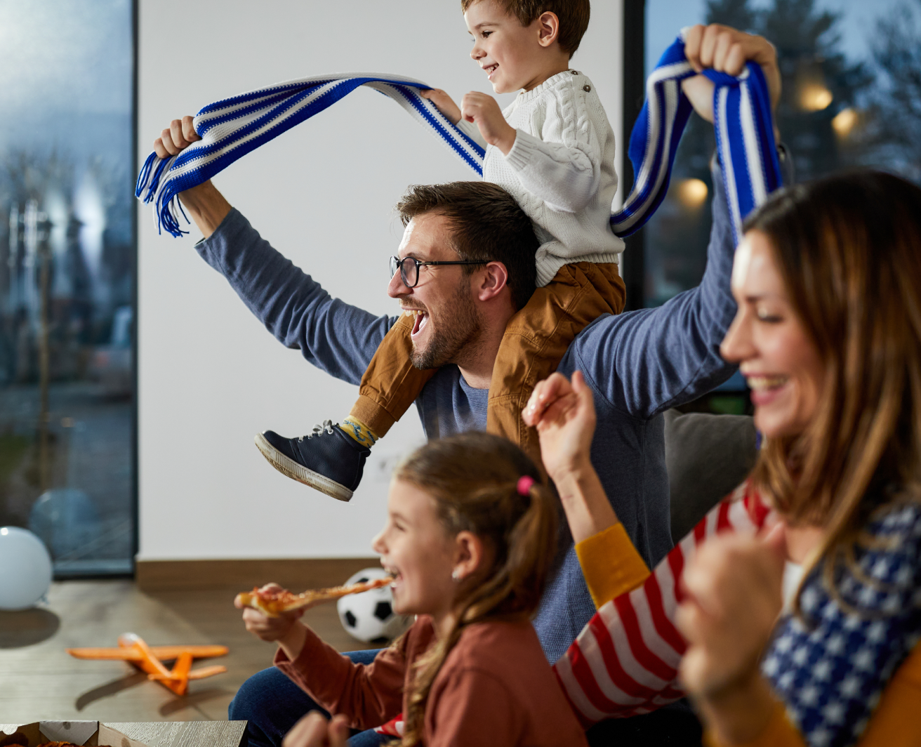 Cilvēki mājas vidē skatās sporta spēli. Priekšplānā ir pieaugušais, kurš uz pleciem tur bērnu ar paceltām rokām, turot zilu un baltu svītrainu šalli. Blakus citas personas, kas arī paceļ rokas, viena tur picas gabalu. Fonā uz grīdas ir rotaļlietas.