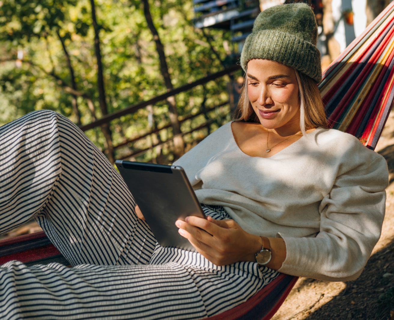 Sieviete atgūlusies krāsainā šūpuļtīklā, turot rokās planšetdatoru. Apģērbā gaišs džemperis, svītrainas bikses un zaļa adīta cepure. Fonā koki un koka margas, kas norāda uz dabas vidi.
