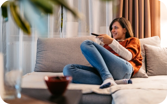Sieviete sēž uz gaiša dīvāna mājīgā istabā, turot TV pulti rokā. Fonā aizkari un dekoratīvi elementi, priekšplānā uz galda novietota bļoda.