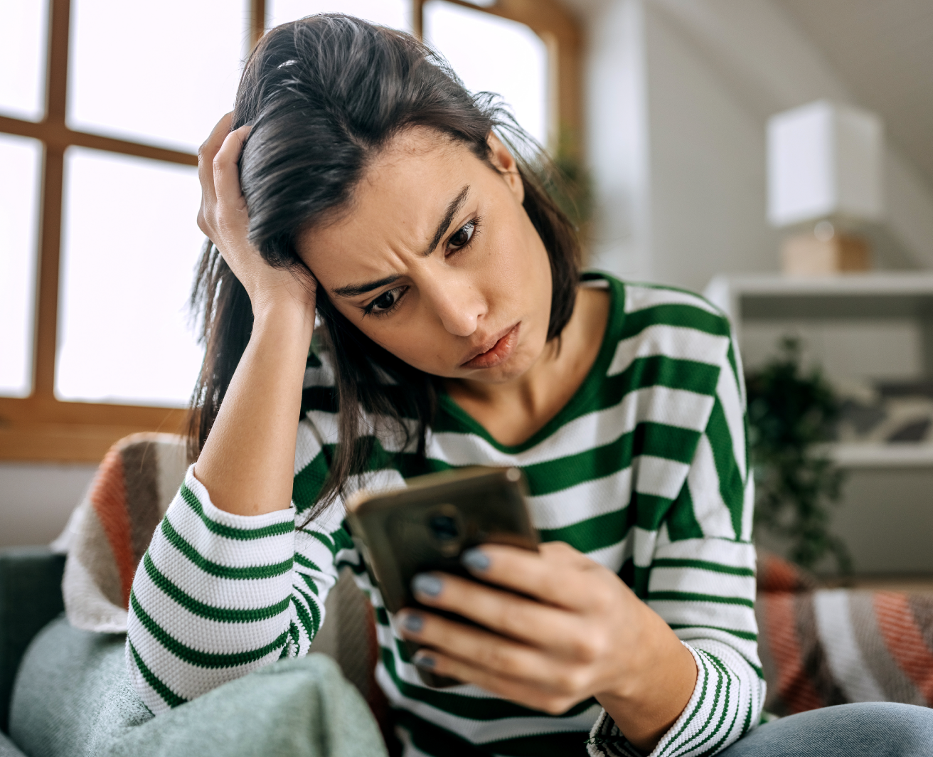 Sieviete skatās savā mobilajā telefonā, turot galvu ar otru roku. Aina satraukuma noskaņu. Fons gaišs.