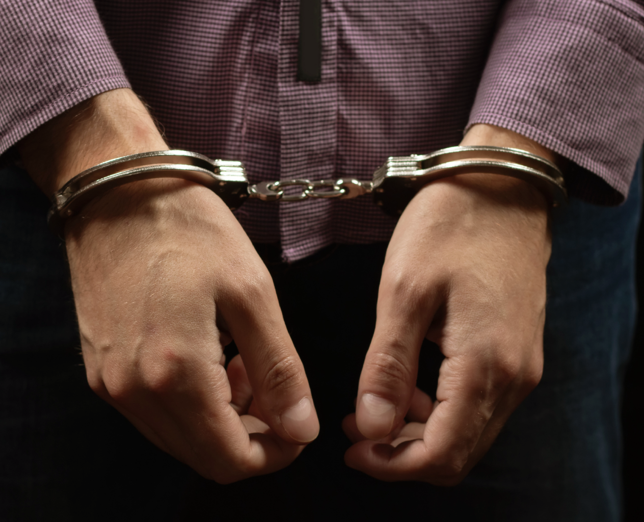 Rokas metāla rokudzelžos, satvertas kopā priekšpusē. Persona tērpusies rūtainā, garroku kreklā, un attēls rada aresta vai aizturēšanas situācijas iespaidu.