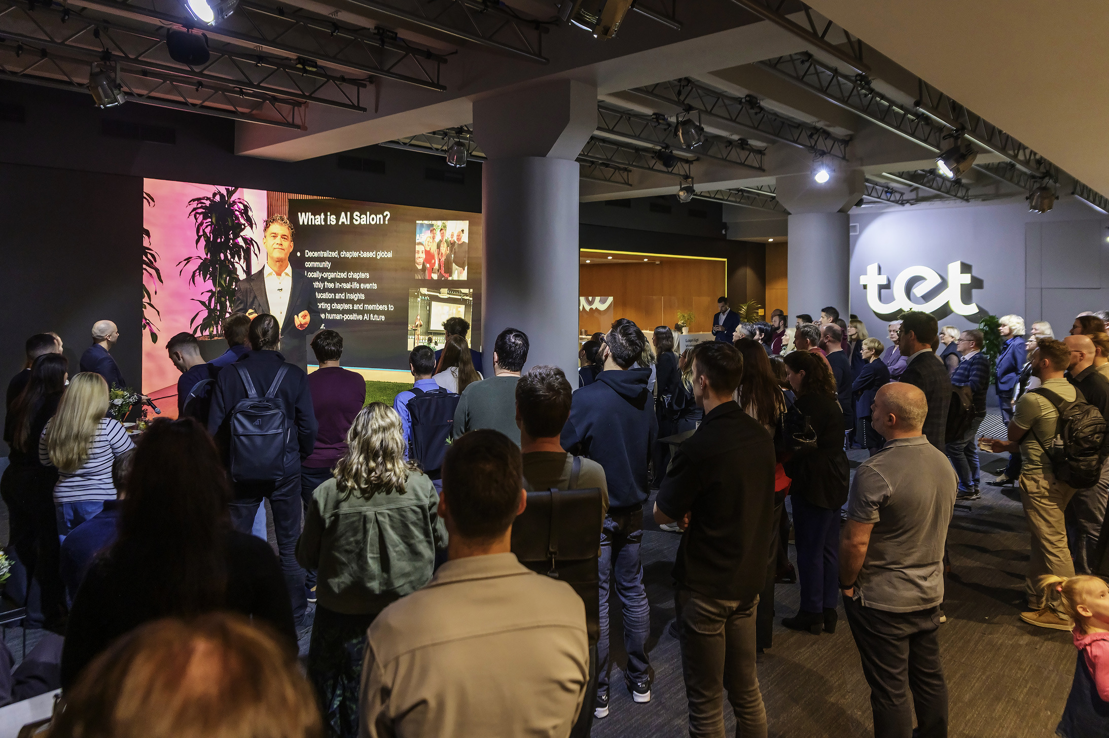 Pasākuma dalībnieki stāv zālē un skatās lekciju par AI Salon uz lielā ekrāna.