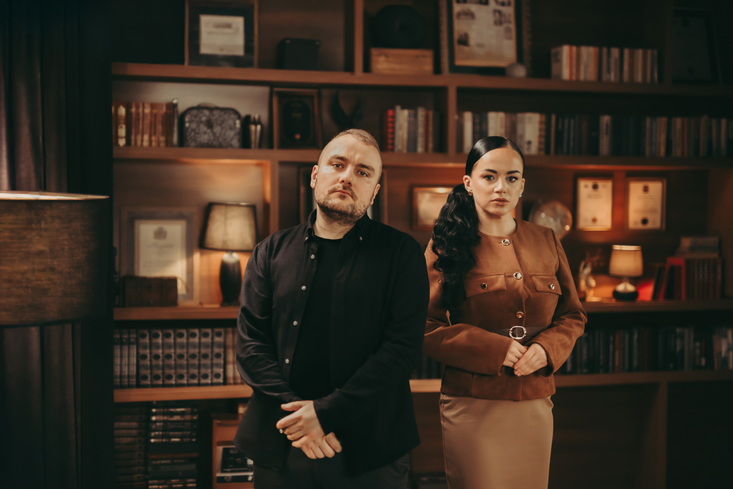 Divi cilvēki kabinetā ar grāmatplauktiem: vīrietis un sieviete stāv, nopietnas sejas.