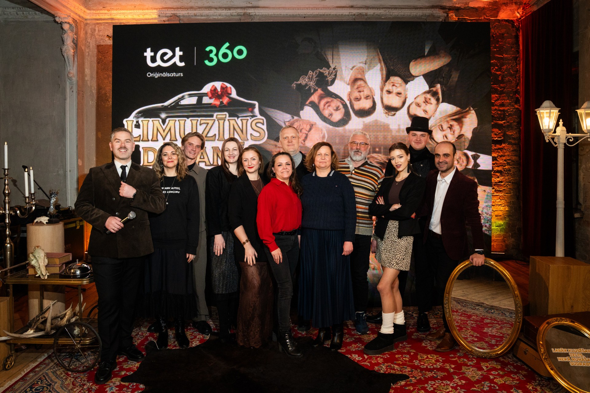 Grupas foto no Tet oriģinālsatura 'Limuzīns dāvanā' prezentācijas pasākuma. Vienpadsmit cilvēki pozē kopā elegantā interjerā ar sarkanu paklāju, fonā redzams seriāla plakāts ar Tet un 360 logo.