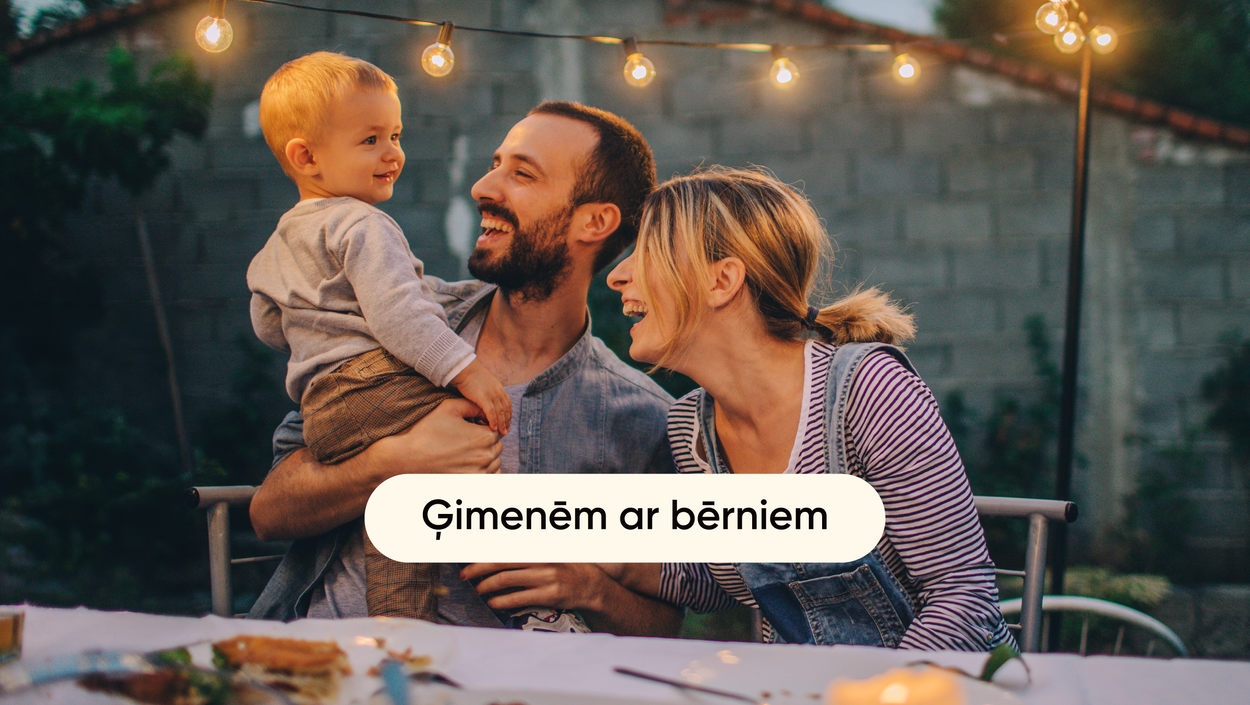 Ģimene ar bērniem sēž pie galda ārā un priecīgi pavada laiku kopā.