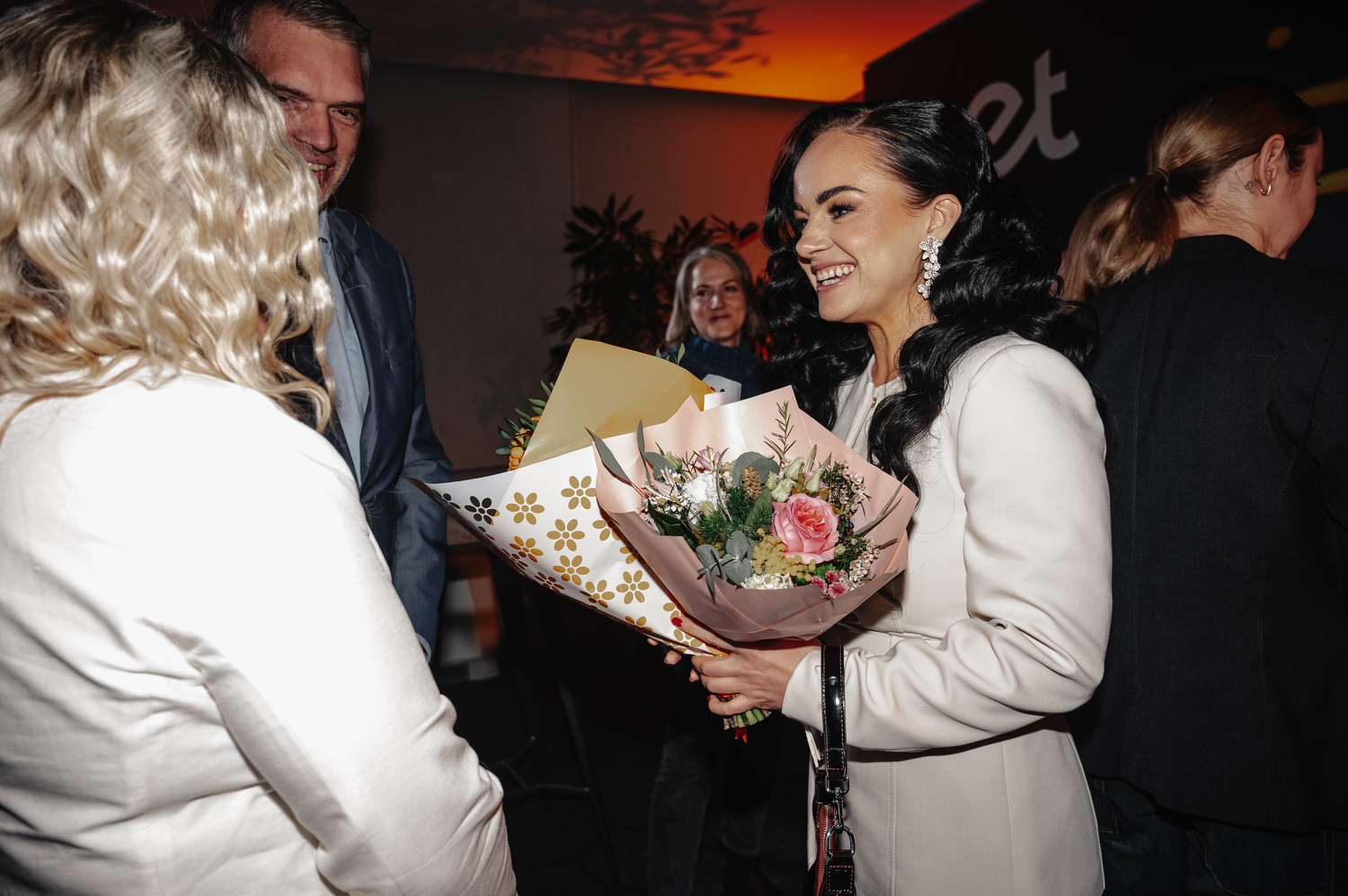 Cilvēku grupa iekštelpās pasākuma laikā. Priekšplānā sieviete ar tumšiem matiem tur vairākus ziedu pušķus dekoratīvā papīrā. Apkārt cilvēki sarunājas.