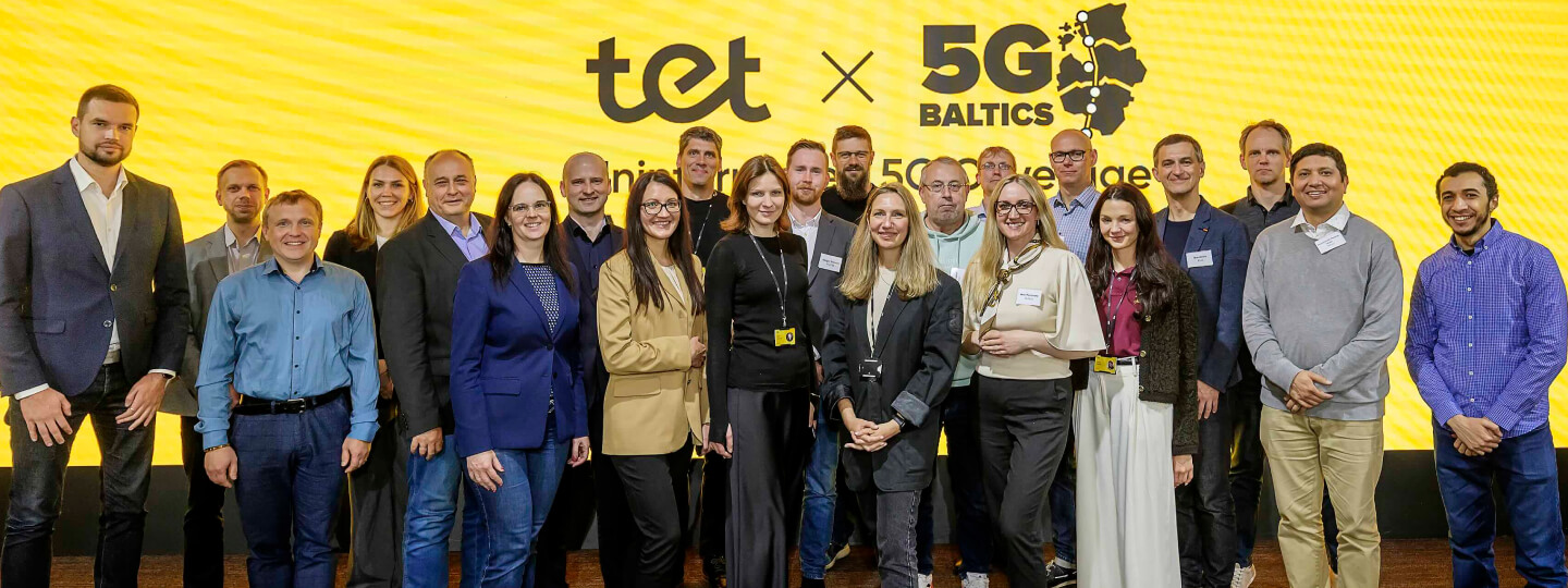 Priekšplānā smaidīgi cilvēki biznesa apģērbā, fonā liels dzeltens ekrāns ar Tet un 5G Baltics logo. Grupas foto no pasākuma.
