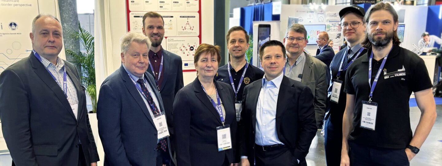 Deviņu cilvēku grupa starptautiskā kiberdrošības forumā pie izstādes stenda ar identifikācijas kartēm ap kaklu; daži ģērbušies uzvalkos, citi – ikdienišķi.