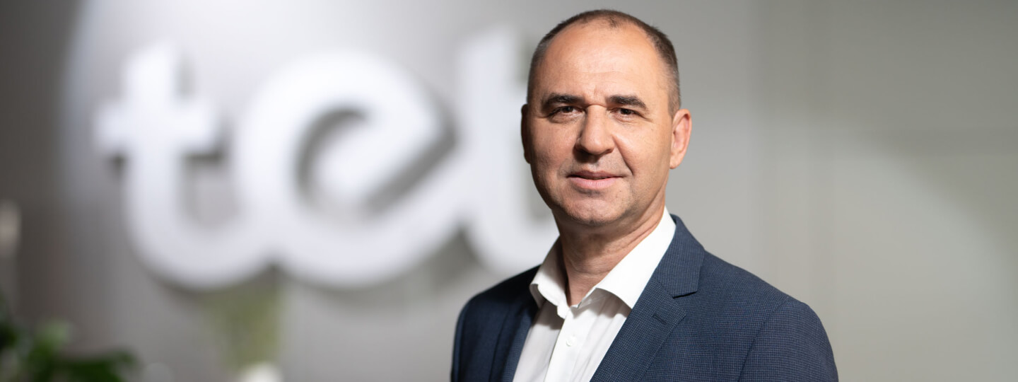 Vīrietis tumšā žaketē un baltā kreklā (Uldis Lībietis, Tet Datu aizsardzības un IT risku nodaļas vadītājs), fonā pelēka siena un Tet logo ar baltiem burtiem