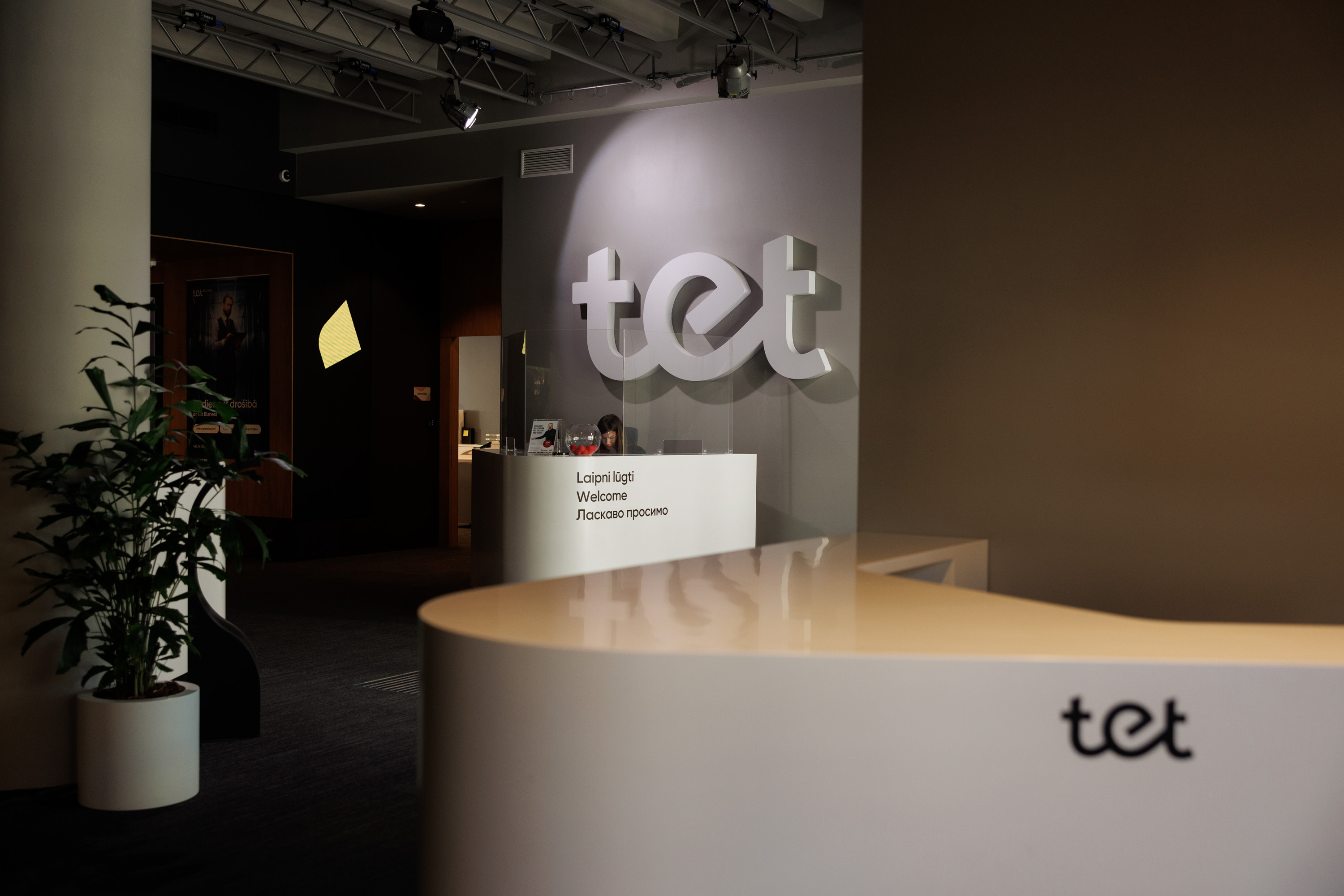 Moderna Tet biroja uzgaidāmā zona ar gaišu, liektu reģistratūras galdu priekšplānā. Fonā siena ar lielu Tet logotipu, telpā ir augi. 