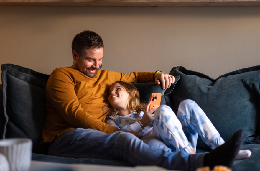 Vīrietis sēž krēslā ar planšetdatoru rokās un bērns pārliecies pāri krēsla malai skatās planšetē