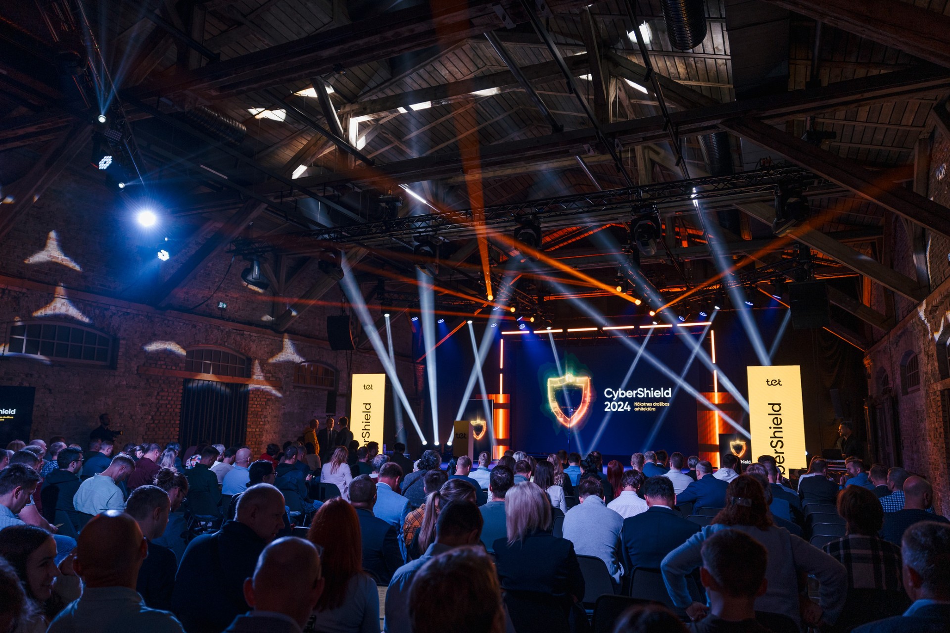 Skatuve ar ‘CyberShield 2024’ ekrānu un gaismu stariem; pilna auditorija industriālā hallē.