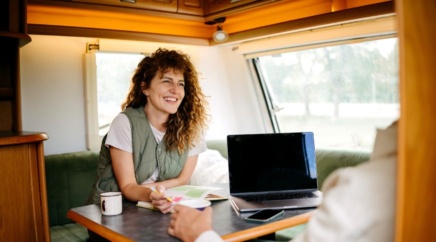 Sieviete kemperī smaidīga sēž pie galda, blakus portatīvais dators