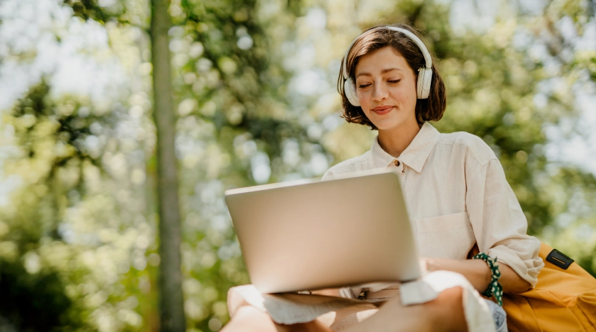 Sieviete smaidīga sēž pargkā uz soliņa, klēpī portatīvais dators