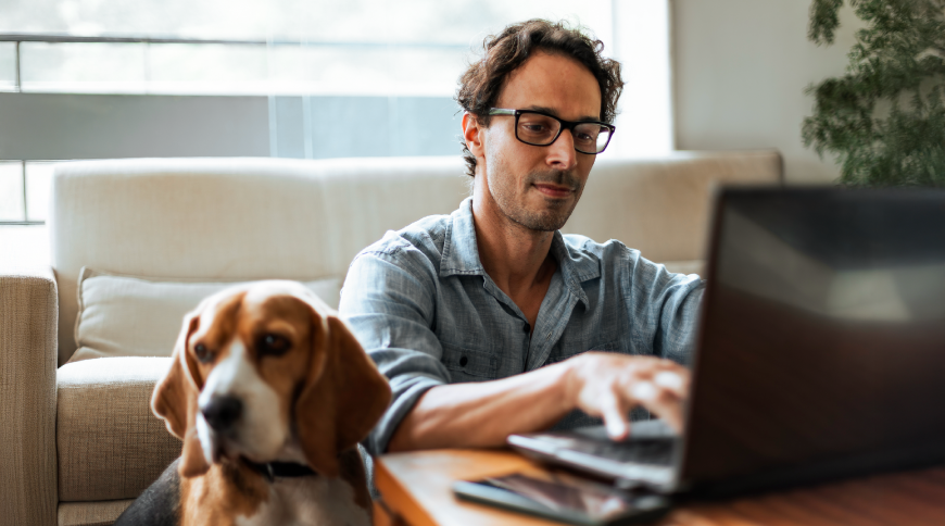 Smaidošs vīrietis strādā pie datora, blakus sēž suns