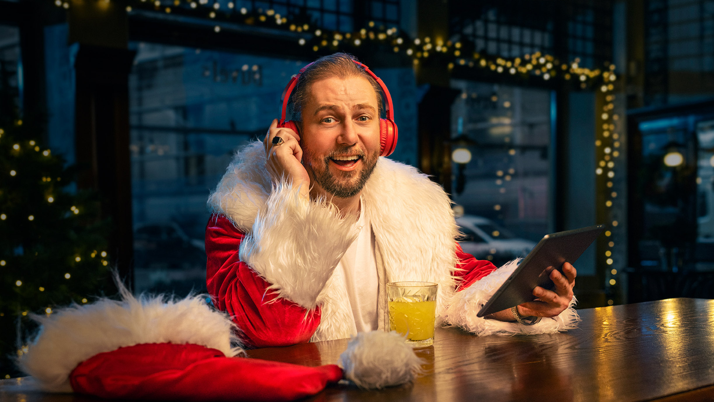Vīrietis Ziemassvētku vecīša tērpā ar sarkanām austiņām pie letes, tur planšeti; fonā svētku gaismiņas.